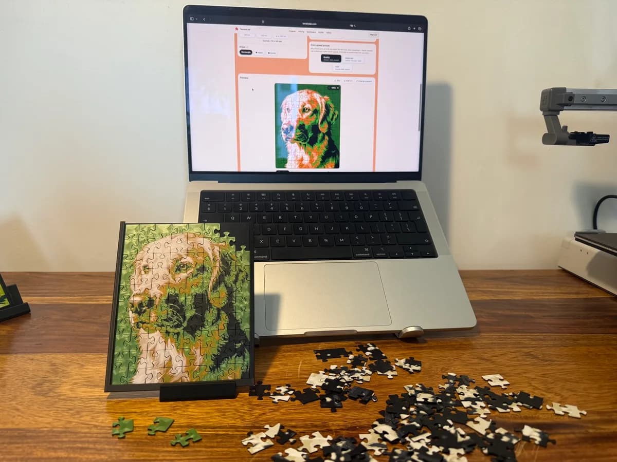 A framed 3D printed photo puzzle of a golden retriever on a wooden desk, in front of a laptop showing the same puzzle in the generator preview, loose pieces scattered nearby