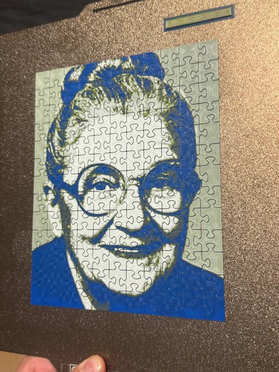 3D printed photo puzzle of a smiling grandmother with glasses, printed in blue, grey and white, on the build plate