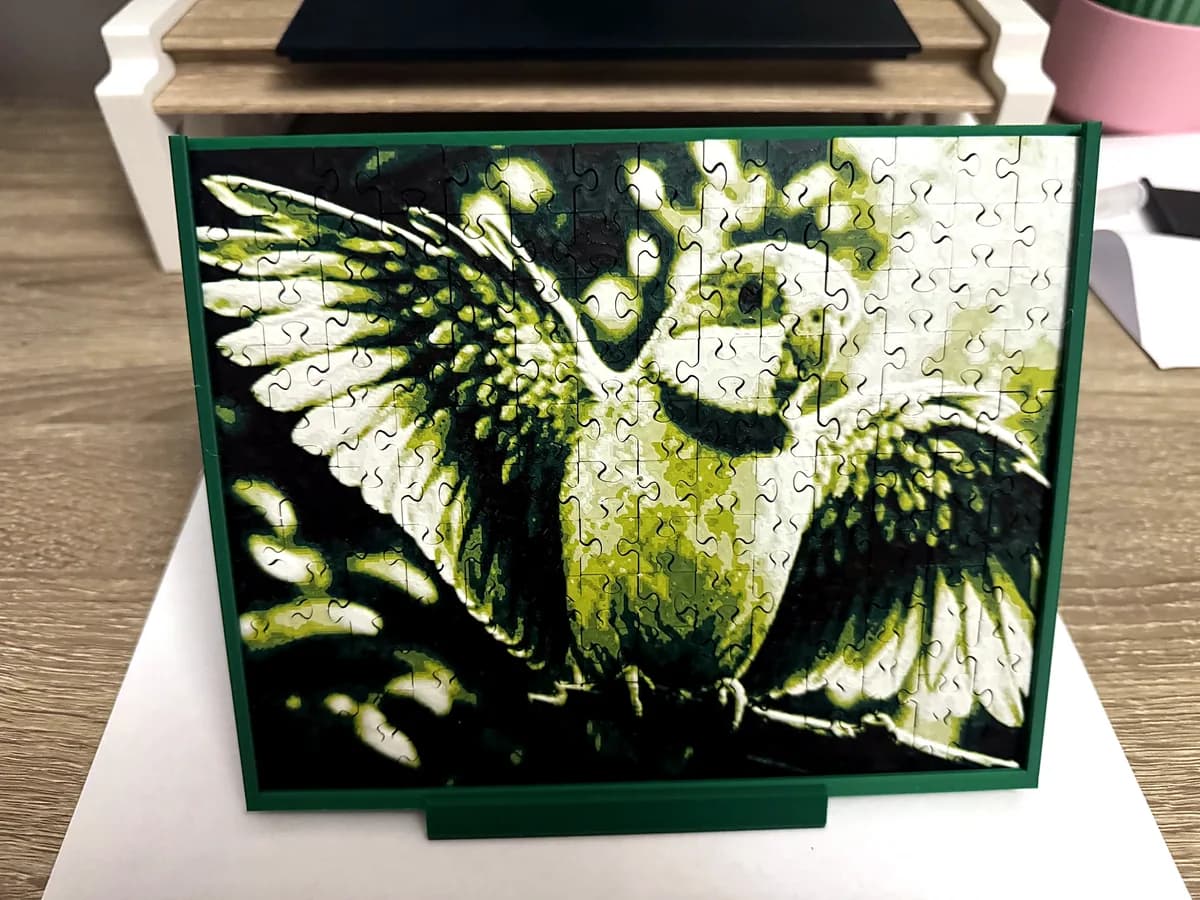 3D printed photo puzzle of a green parrot in a green frame with a stand, on a wooden desk